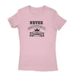 Image of a Light Pink t-shirt with a white text graphic that reads 'NEVER UNDERESTIMATE THE POWER' with a crown symbol above the text, promoting a message of empowerment and resilience.